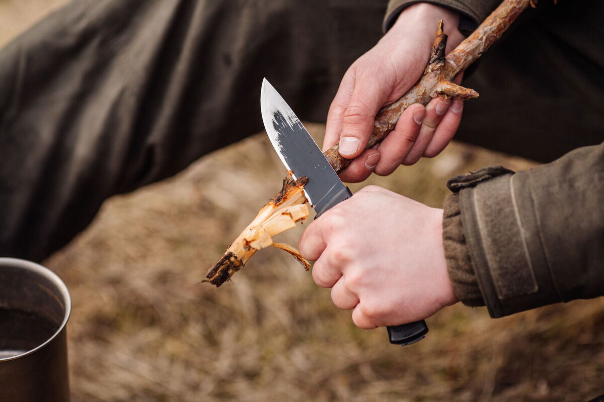 A close-up of a man's hands. He is holding a small hand knife to whittle down the end of a stick.