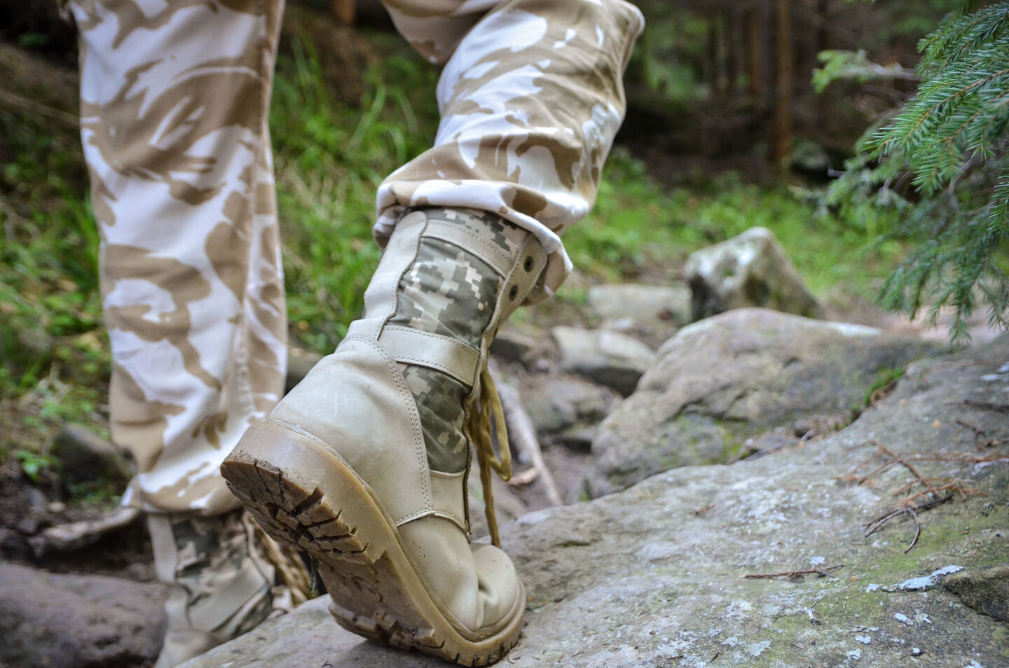 A soldier wearing military-style trekking shoes and pants, walking on a rocky path in a green forest.