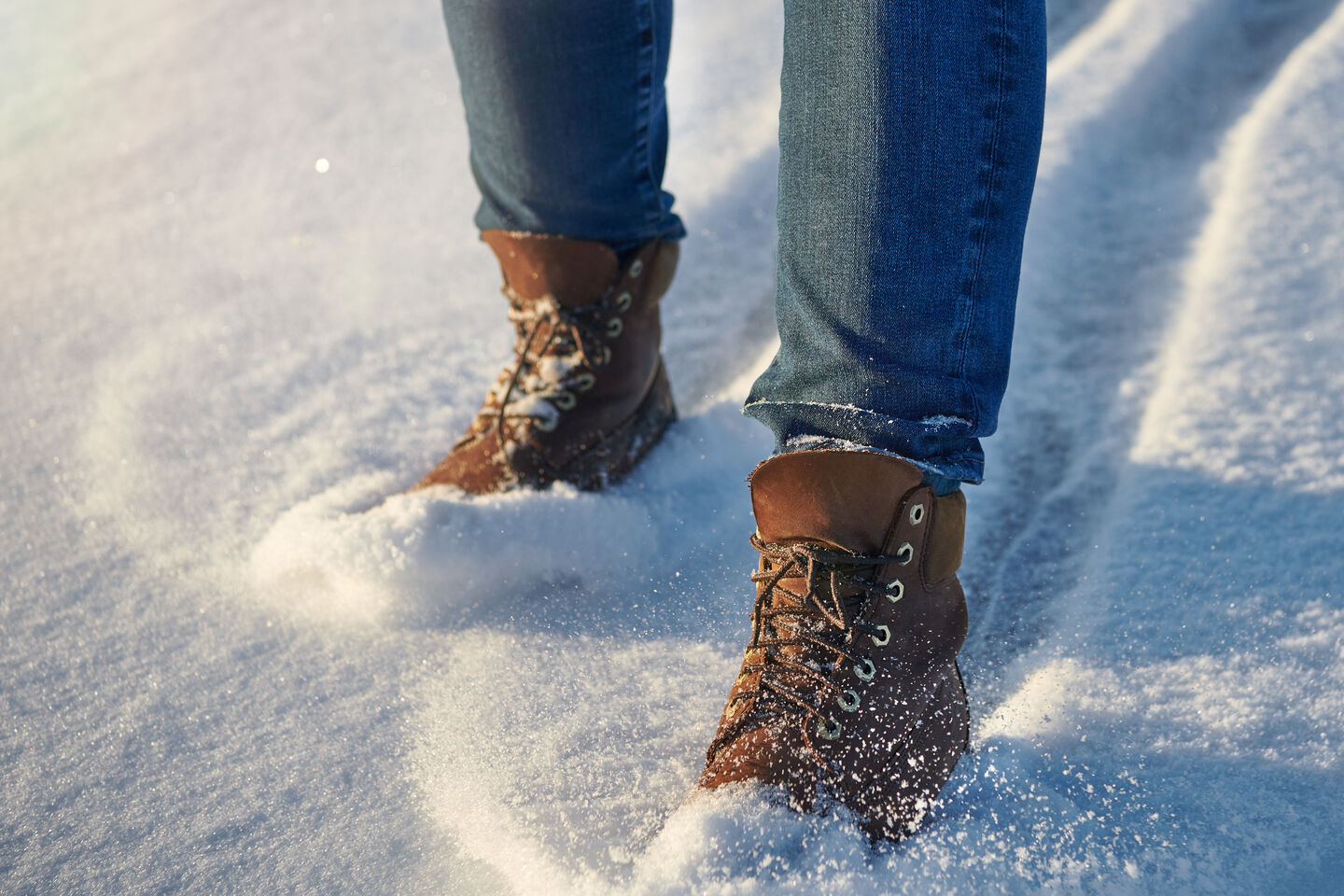 A close-up of a person's legs in brown leather boots and jeans, walking in fresh snow and leaving a trail in their wake.